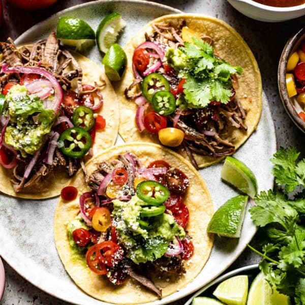 Brisket Tacos served over corn tortillas on a plate with toppings like guacamole, jalapeno, and tomatoes.