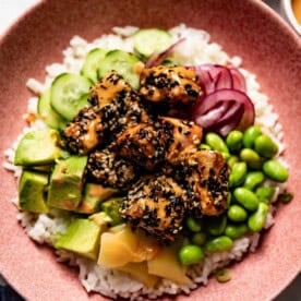 A bowl of rice topped with sesame-crusted salmon, avocado, edamame, and pickled onions.