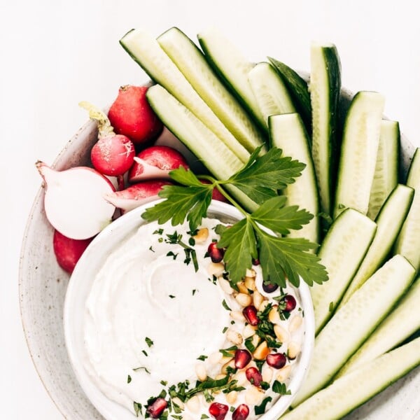 A bowl of tahini yogurt sauce surrounded with cucumbers and radishes is photographed from the top view.