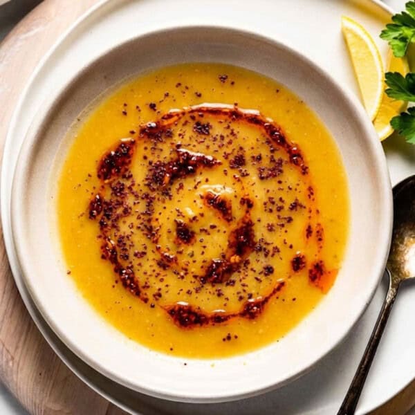 A close-up, top-down view of a bowl of smooth Turkish red lentil soup, drizzled with a swirl of red chili butter (pul biber yağı) and sprinkled with dark red Aleppo pepper flakes, served with a side of lemon and parsley.